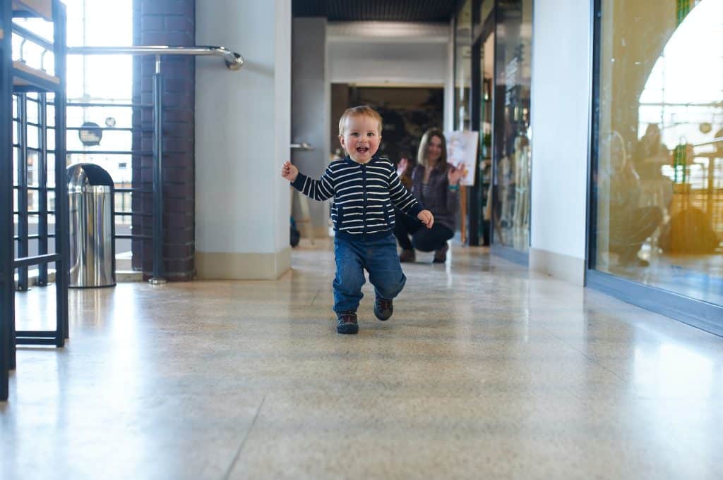 Toddler running in the shopping center with his mom on background.