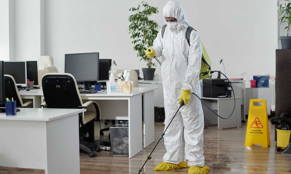 Male worker of cleaning service in hazmat suit disinfecting floor in office