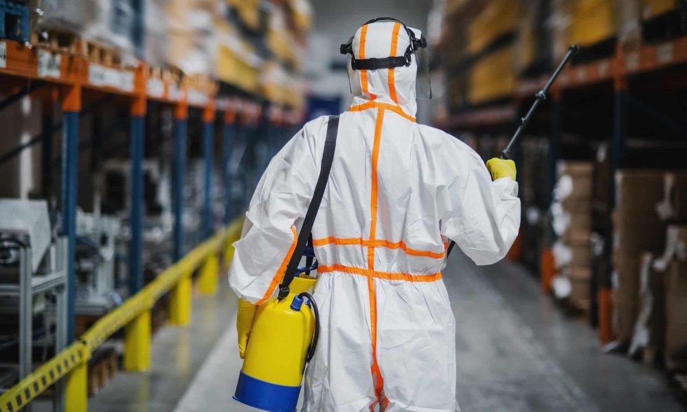 Rear view of man worker with protective suit disinfecting industrial factory with spray gun
