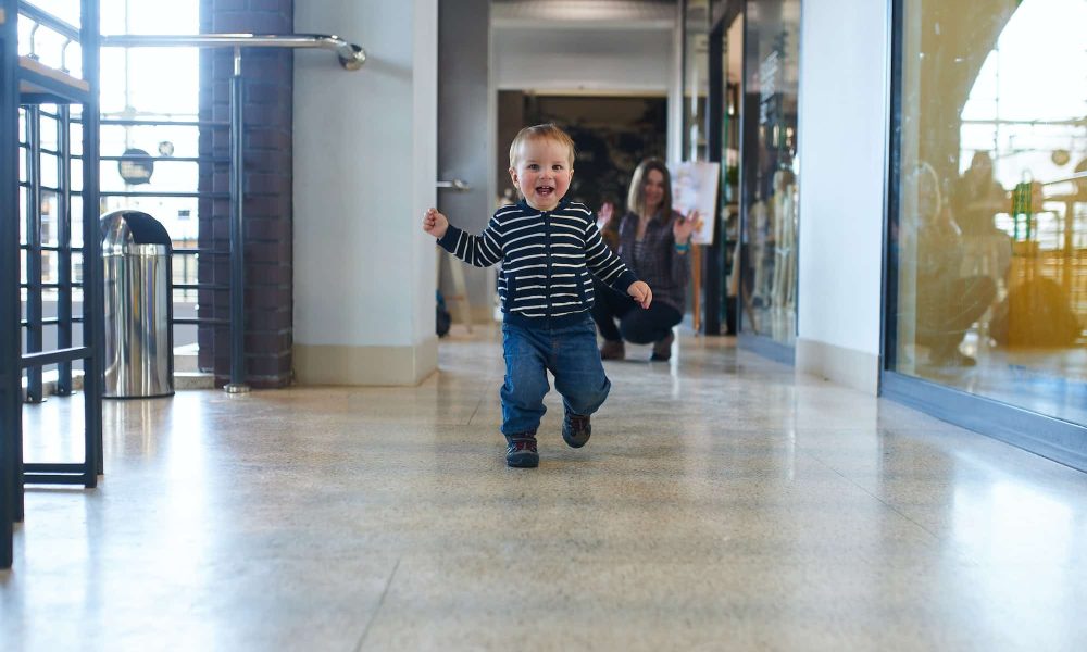 Toddler running in the shopping center with his mom on background.